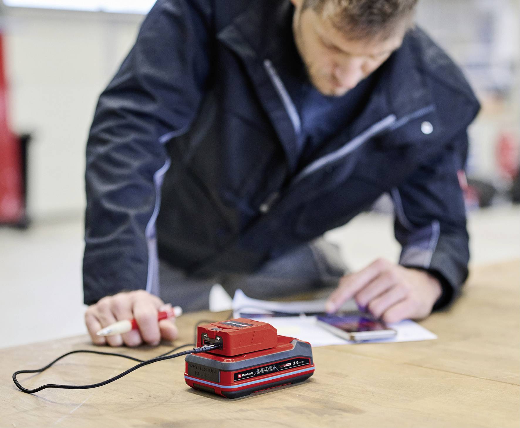 Une personne en arrière-plan écrit sur du papier. Au premier plan, un outil électrique rouge est posé sur une table.