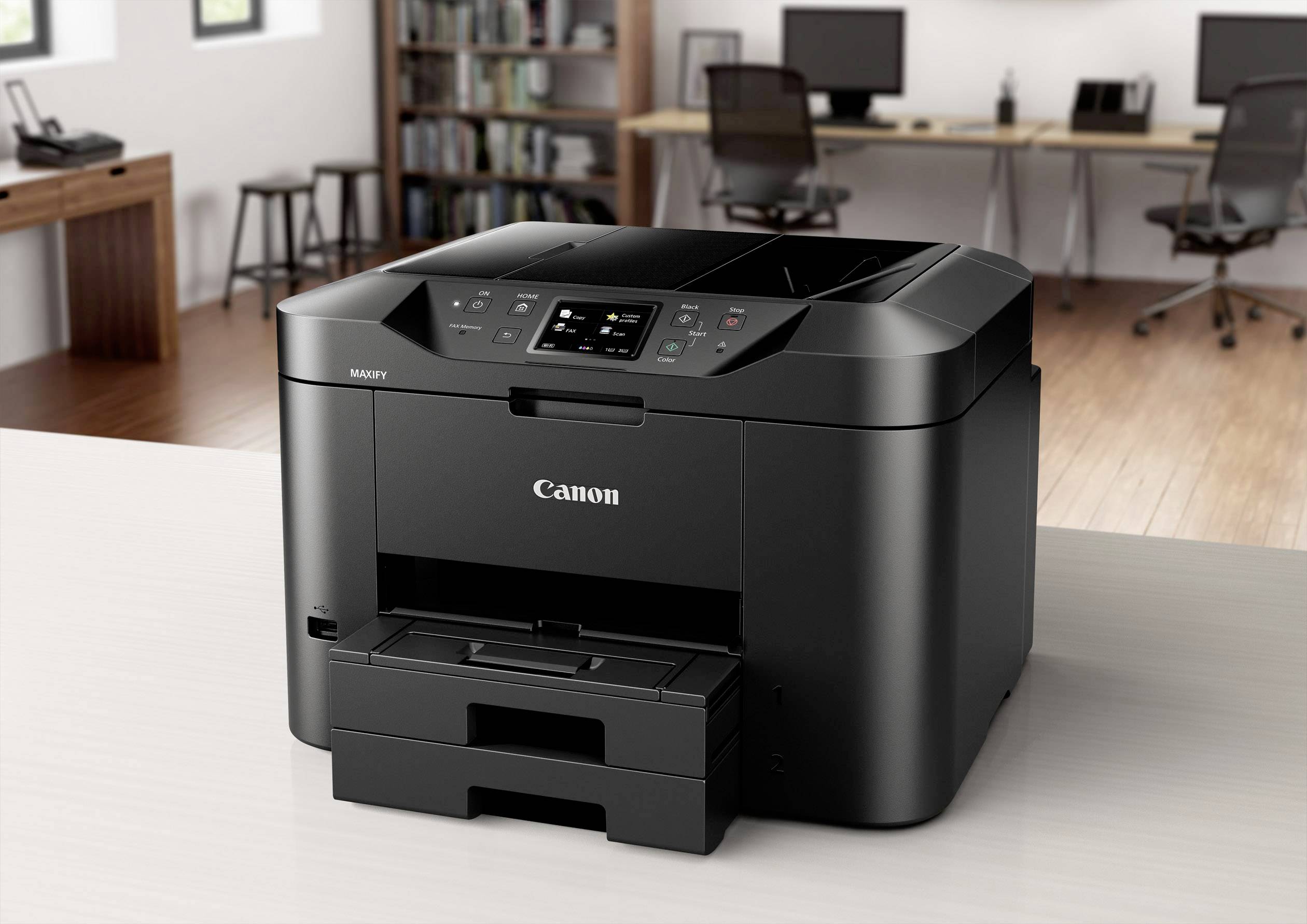 Un imprimante noir Canon est posée sur un bureau dans un bureau moderne avec des étagères et des ordinateurs en arrière-plan.