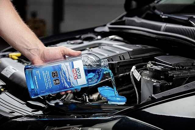 Une personne verse du liquide de lave-glace bleu dans le réservoir ouvert d'une voiture. Le compartiment moteur est visible.