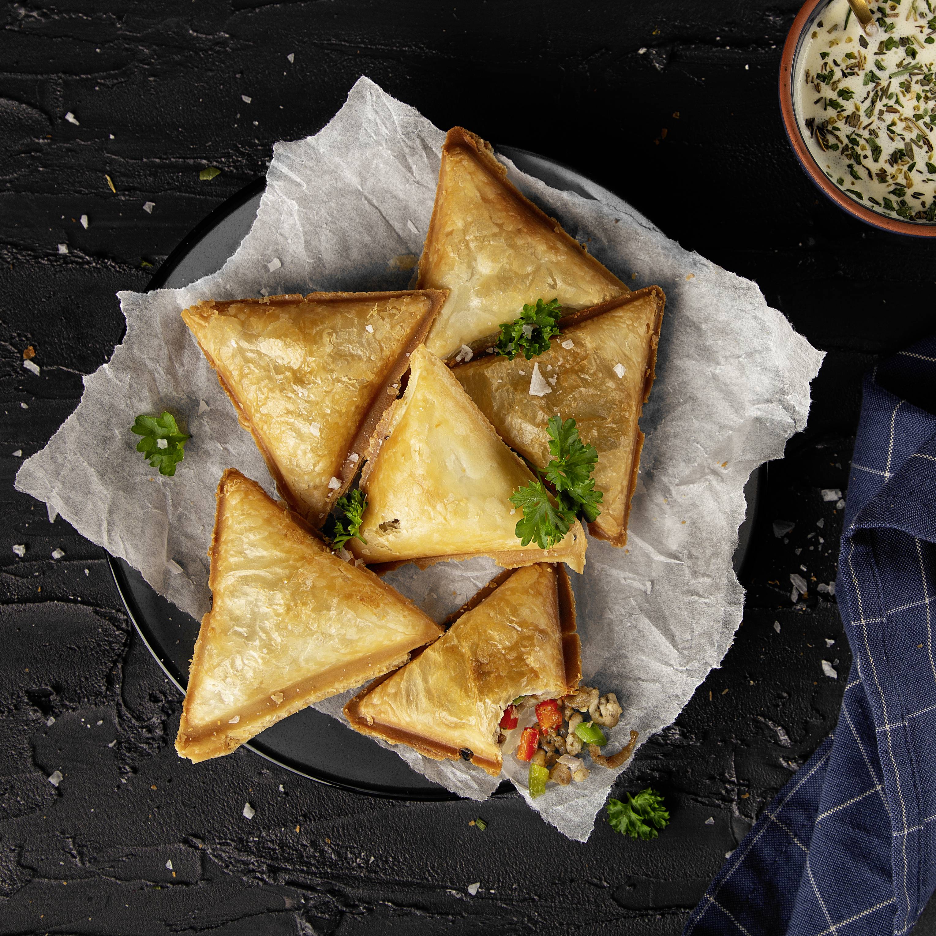 Chaussons triangulaires à la croûte dorée sur du papier sulfurisé. Garnis de légumes, accompagnés d'un bol de sauce au yaourt.