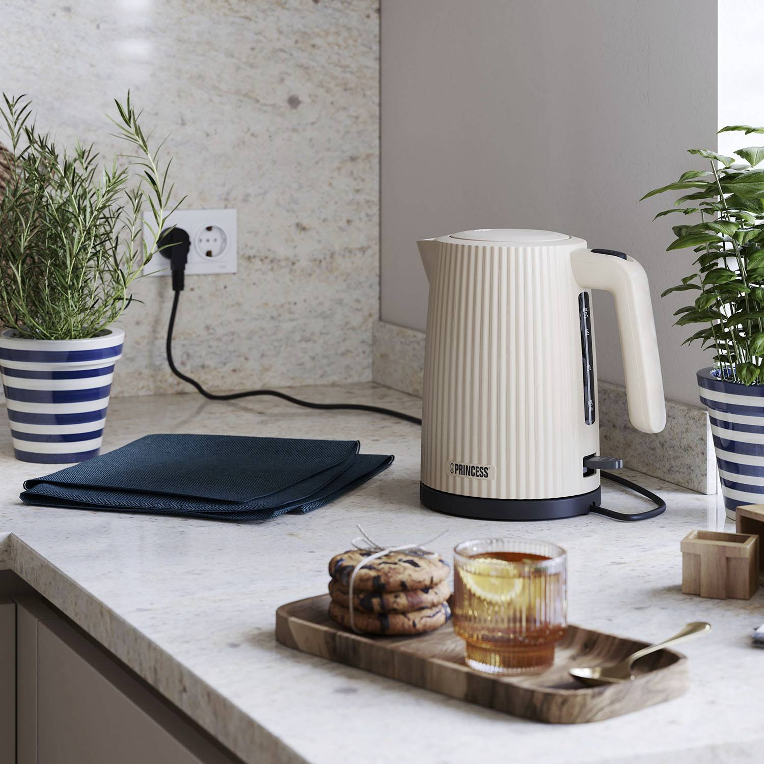 Une bouilloire est posée sur un plan de travail de cuisine à côté de plantes et d'une tasse contenant une boisson et des biscuits sur une planche en bois.