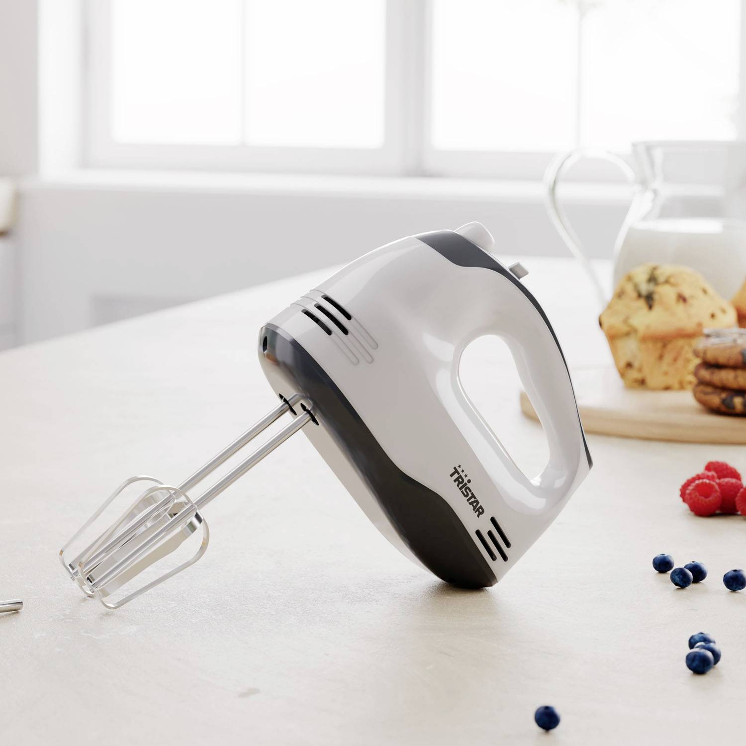 Un batteur électrique sur un plan de travail de cuisine à côté d'un verre de lait et de quelques biscuits. La lumière du jour entre par la fenêtre.