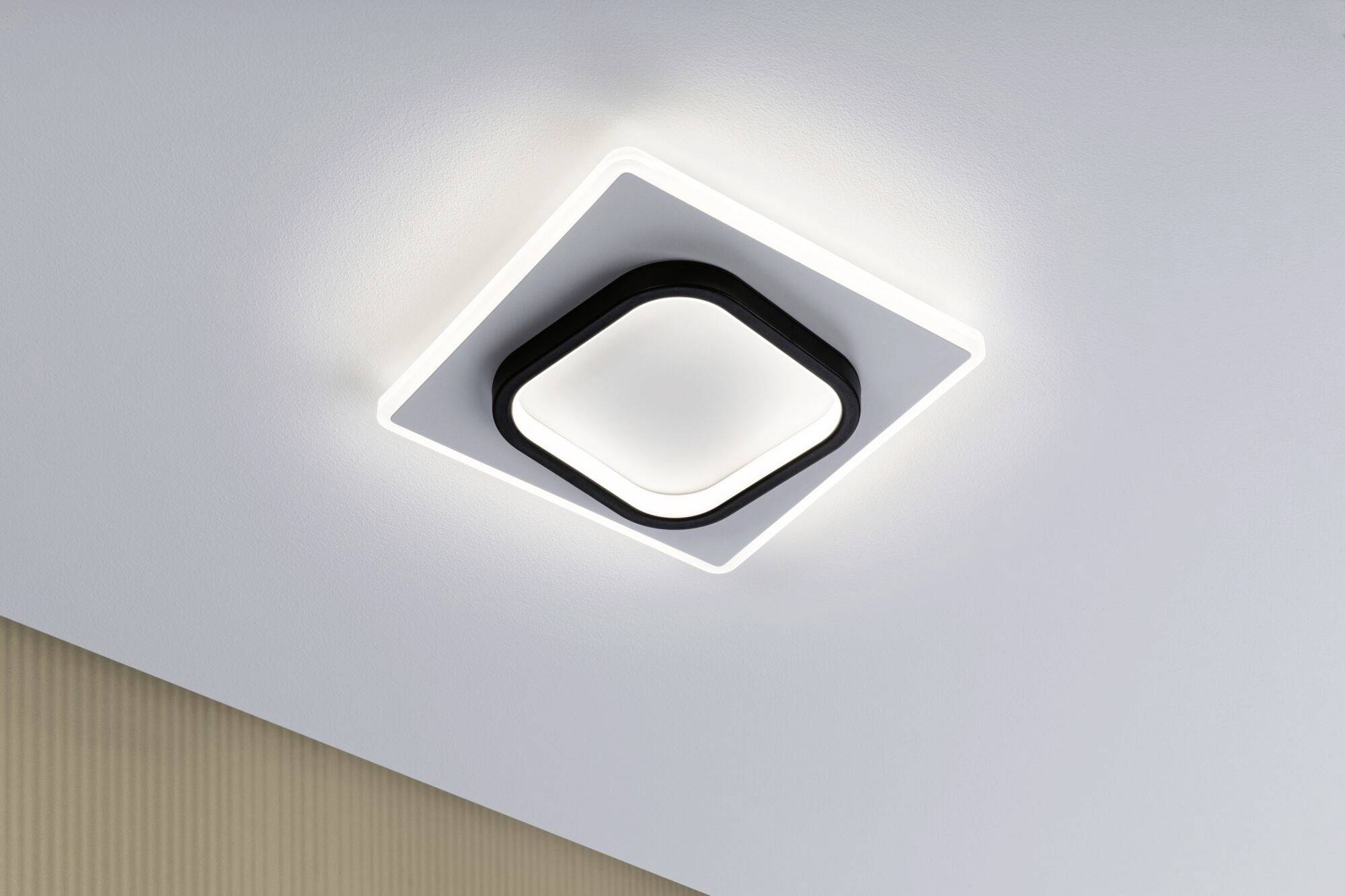 Luminaire de plafond carré moderne avec cadre noir, éclairé de l'intérieur, installé sur un plafond clair dans une pièce.