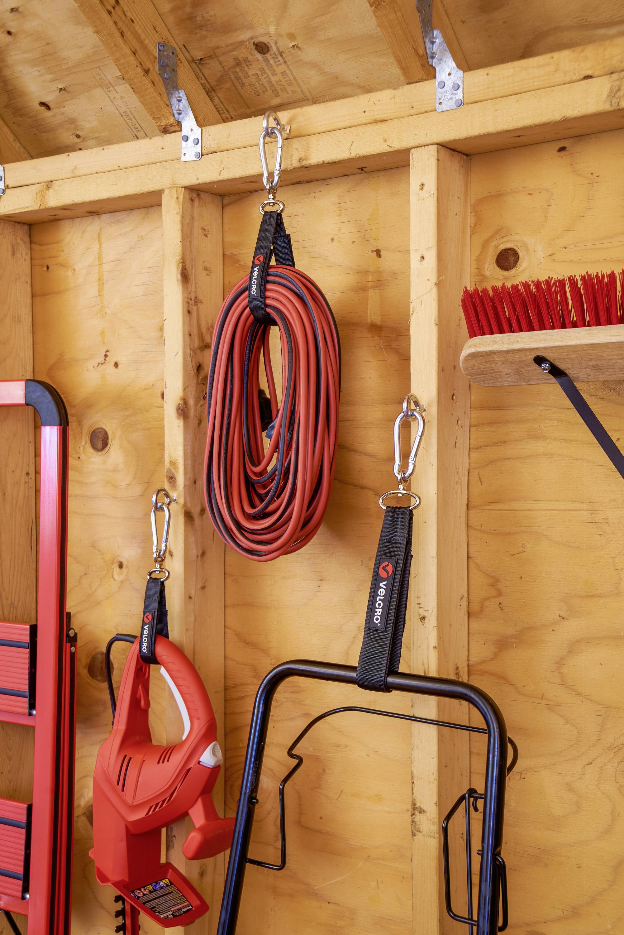Dans un cabanon en bois, différents outils sont accrochés au mur : un câble de rallonge rouge, un souffleur de feuilles et un balai.