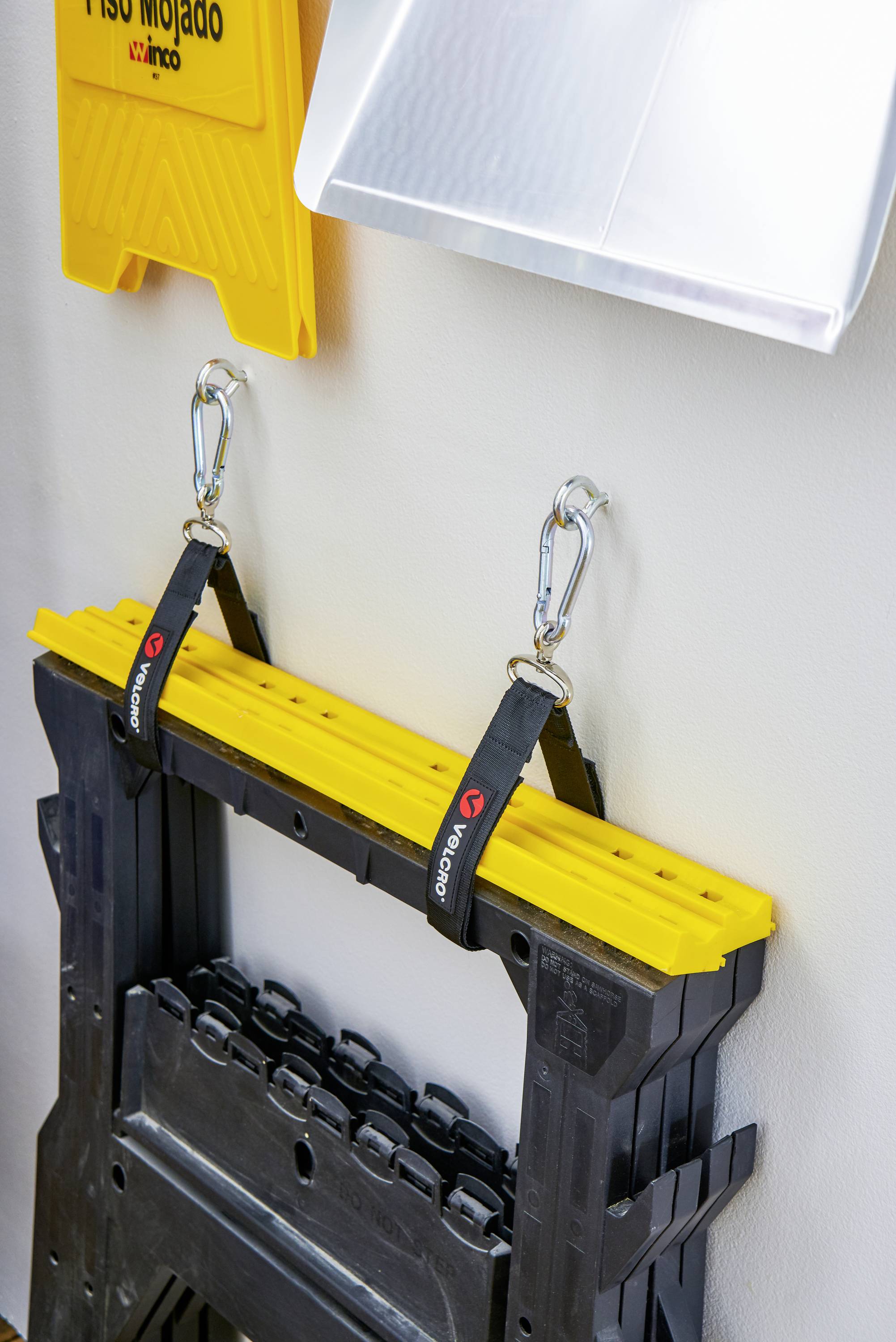 Un établi pliant noir avec un plateau jaune est suspendu à deux crochets au mur. Au-dessus se trouve un panneau indiquant 'Piso Mojado'.
