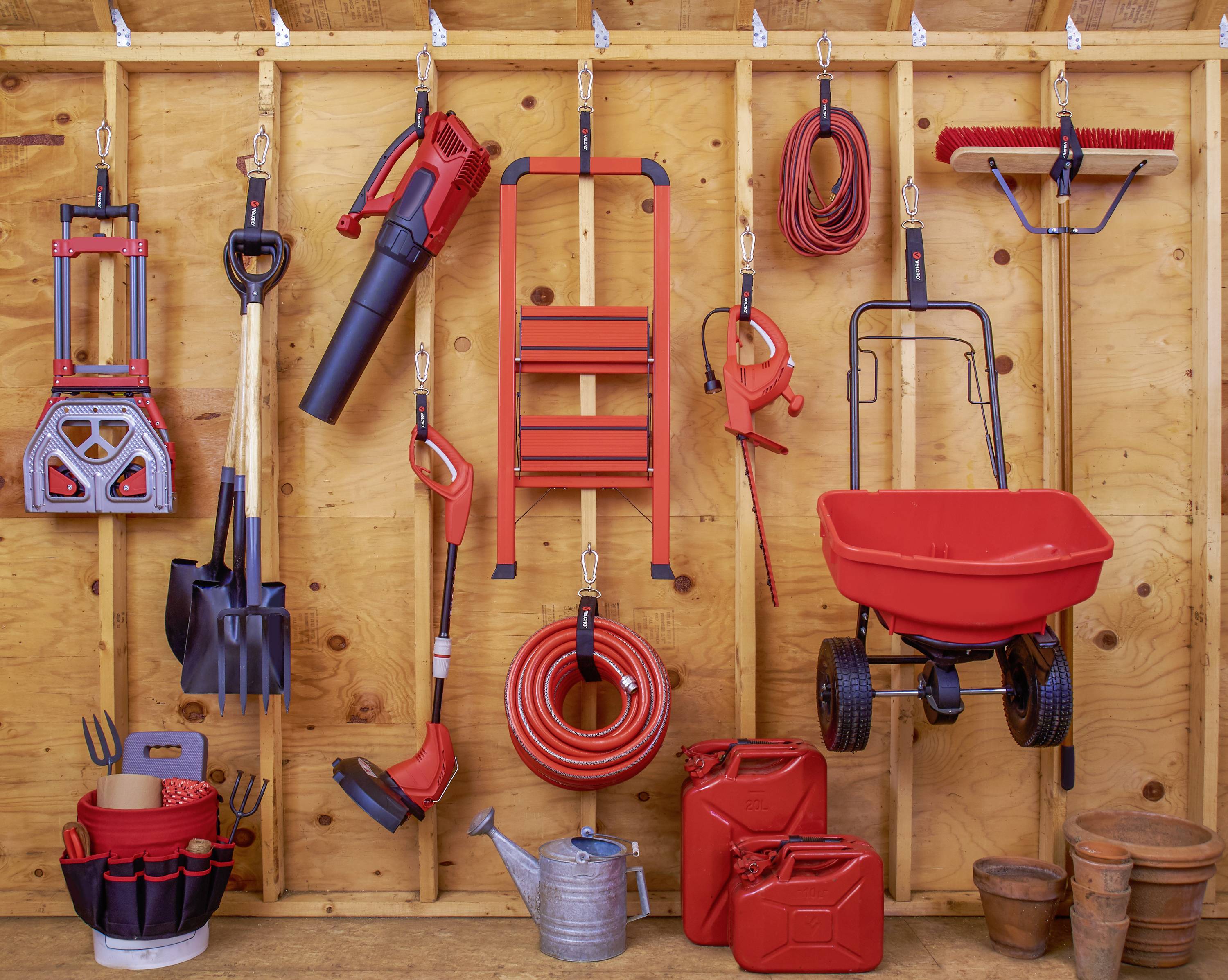 Divers outils de jardin rouges sont suspendus à un mur en bois, notamment des pelles, une brouette et un tuyau d'arrosage.