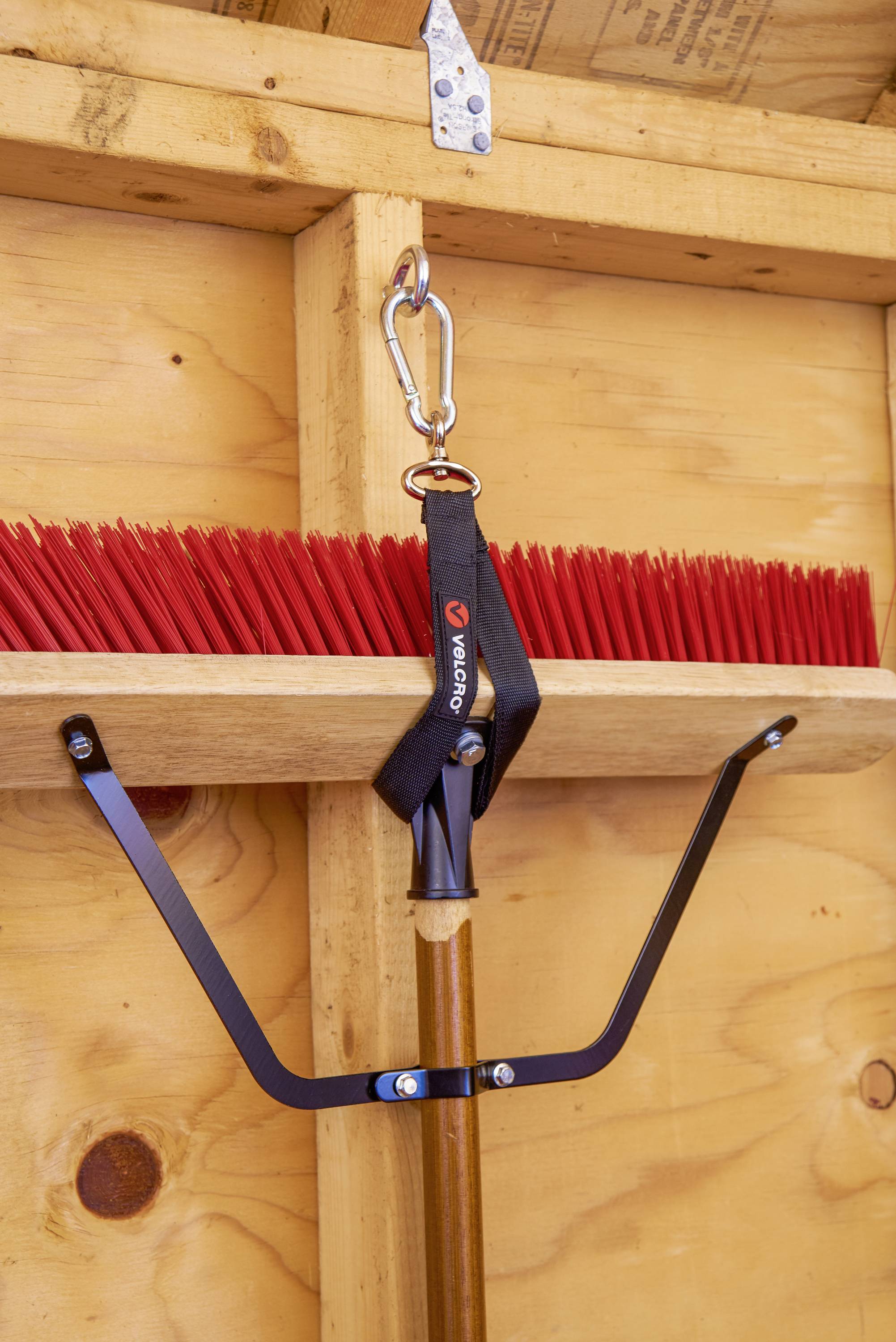 Un balai aux poils rouges est accroché au mur d'un cabanon en bois, maintenu par un support noir avec un mousqueton.