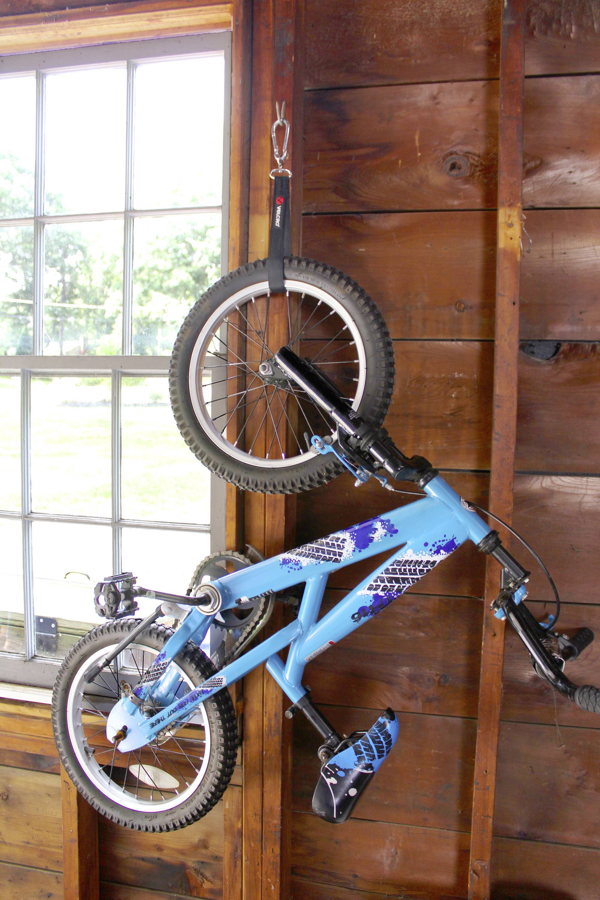 Un vélo d'enfant bleu est suspendu par une sangle à un mur en bois à côté d'une fenêtre.