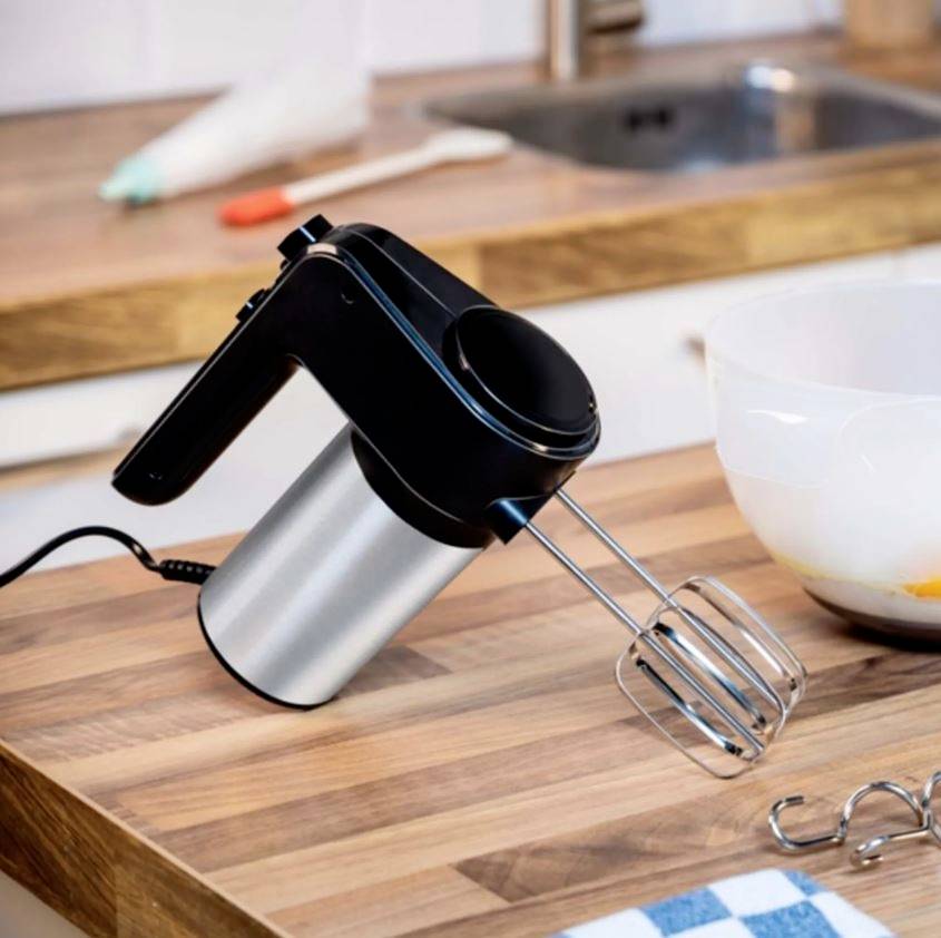 Robot de cuisine sur un plan de travail en bois, avec des fouets installés. À côté, un bol blanc, prêt pour la pâtisserie ou la cuisine.