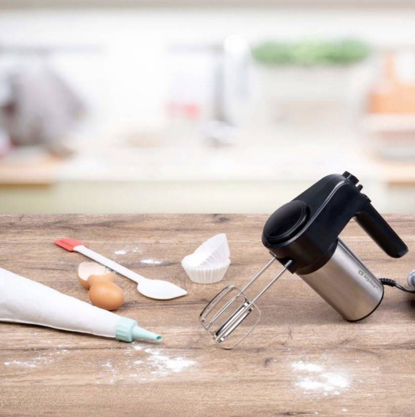 Un batteur électrique est posé sur une table en bois à côté d'une poche à douille, d'un œuf, d'une cuillère à pâtisserie et d'un moule en papier dans une cuisine.
