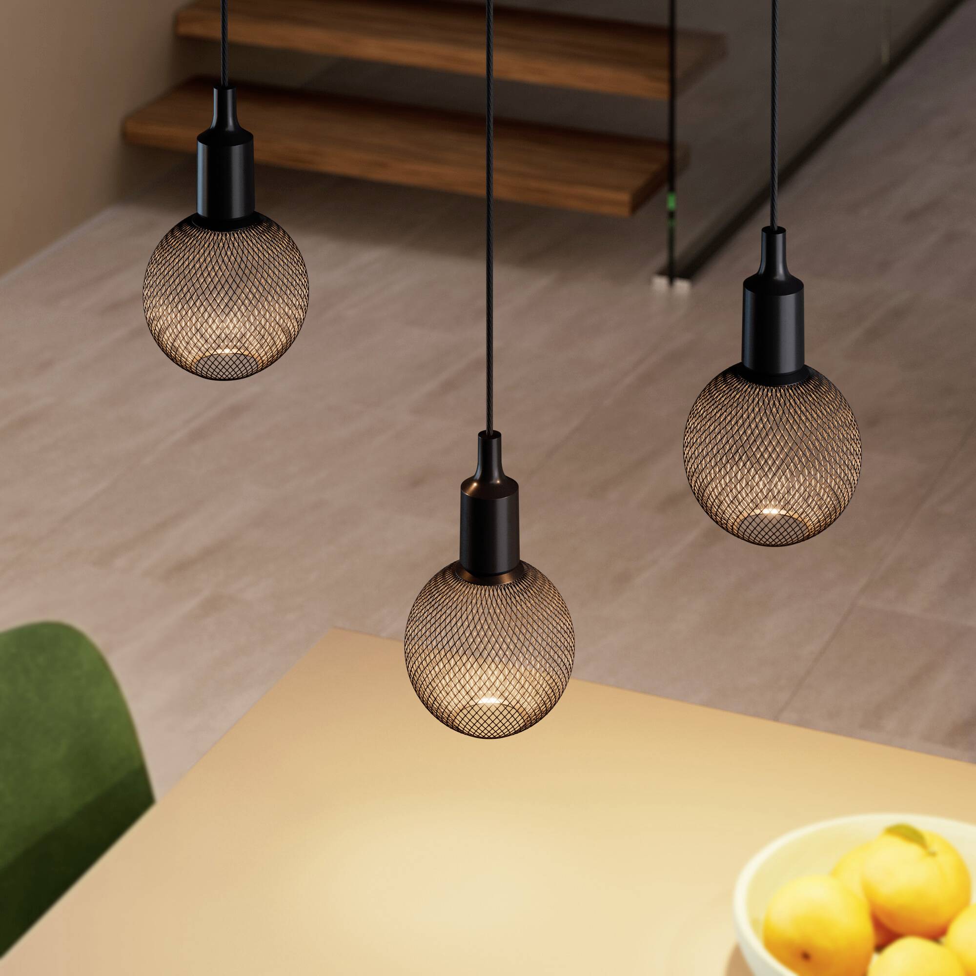 Trois lampes suspendues noires avec des abat-jour en grillage, brillant au-dessus d'une table. Une cage d'escalier en bois en arrière-plan.