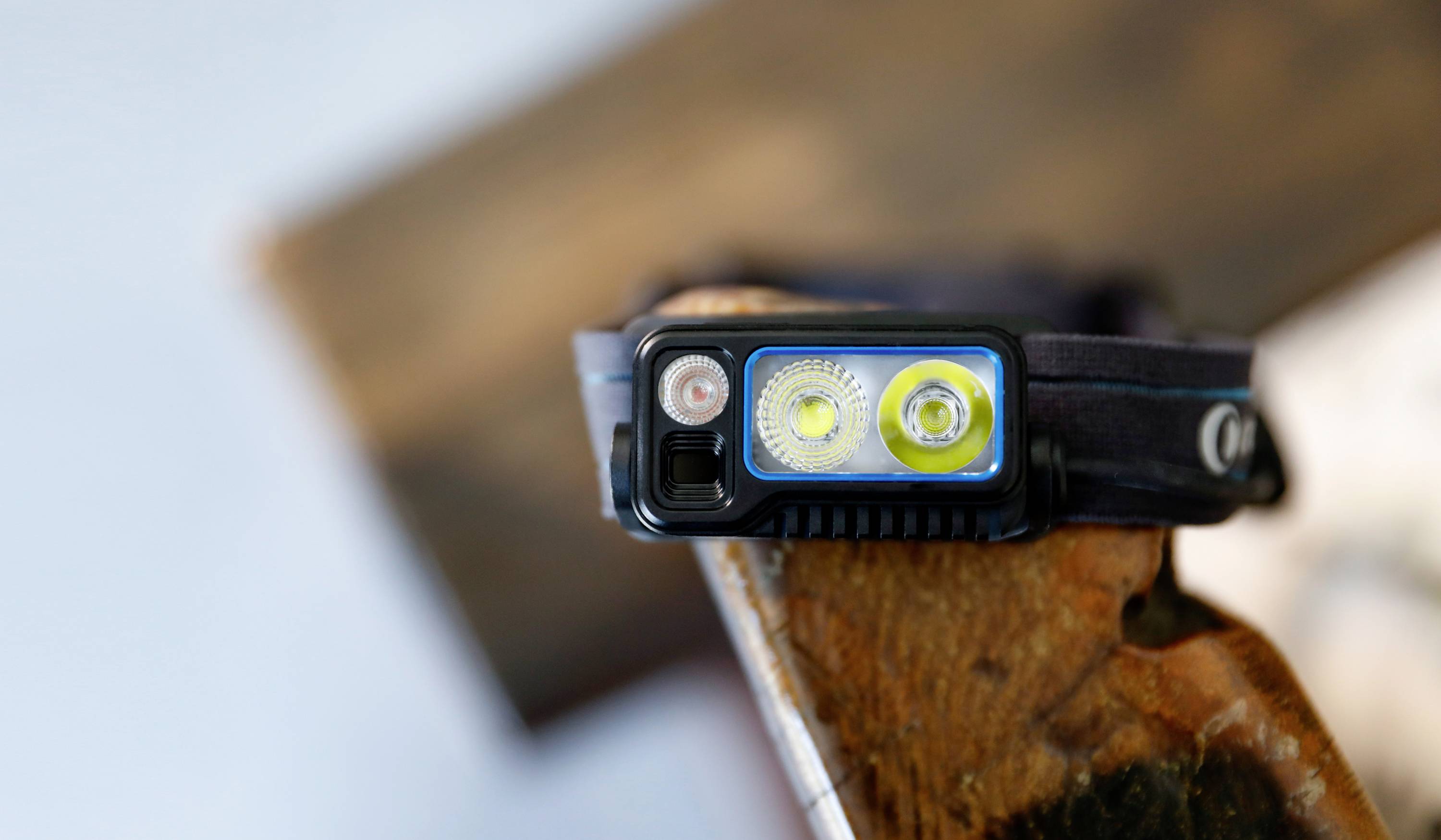 Une lampe frontale noire avec trois sources lumineuses LED est posée sur un morceau de bois. Elle semble être résistante aux chocs et robuste pour les activités de plein air.