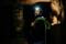 Un homme portant un casque avec lampe frontale et une corde d'escalade regarde vers le haut dans une grotte, baigné d'une lumière chaleureuse. L'environnement est sombre et rustique.