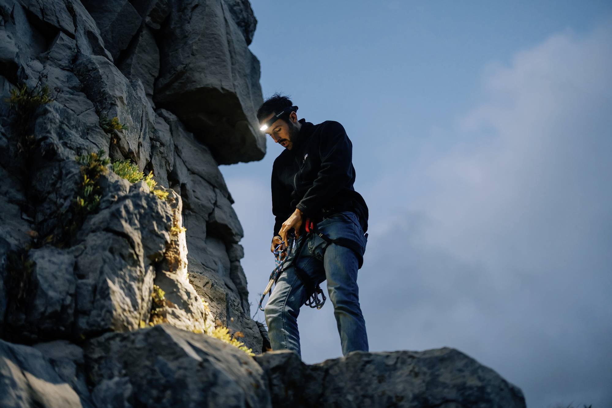 Un grimpeur avec un lampadaire frontal se tient sur un rocher, faisant passer une corde à travers un dispositif d'assurage. L'arrière-plan montre un ciel nuageux au crépuscule.