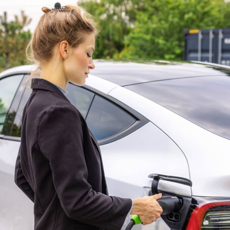 Une femme charge une voiture électrique en branchant le câble de charge sur le véhicule. Des arbres sont visibles en arrière-plan.