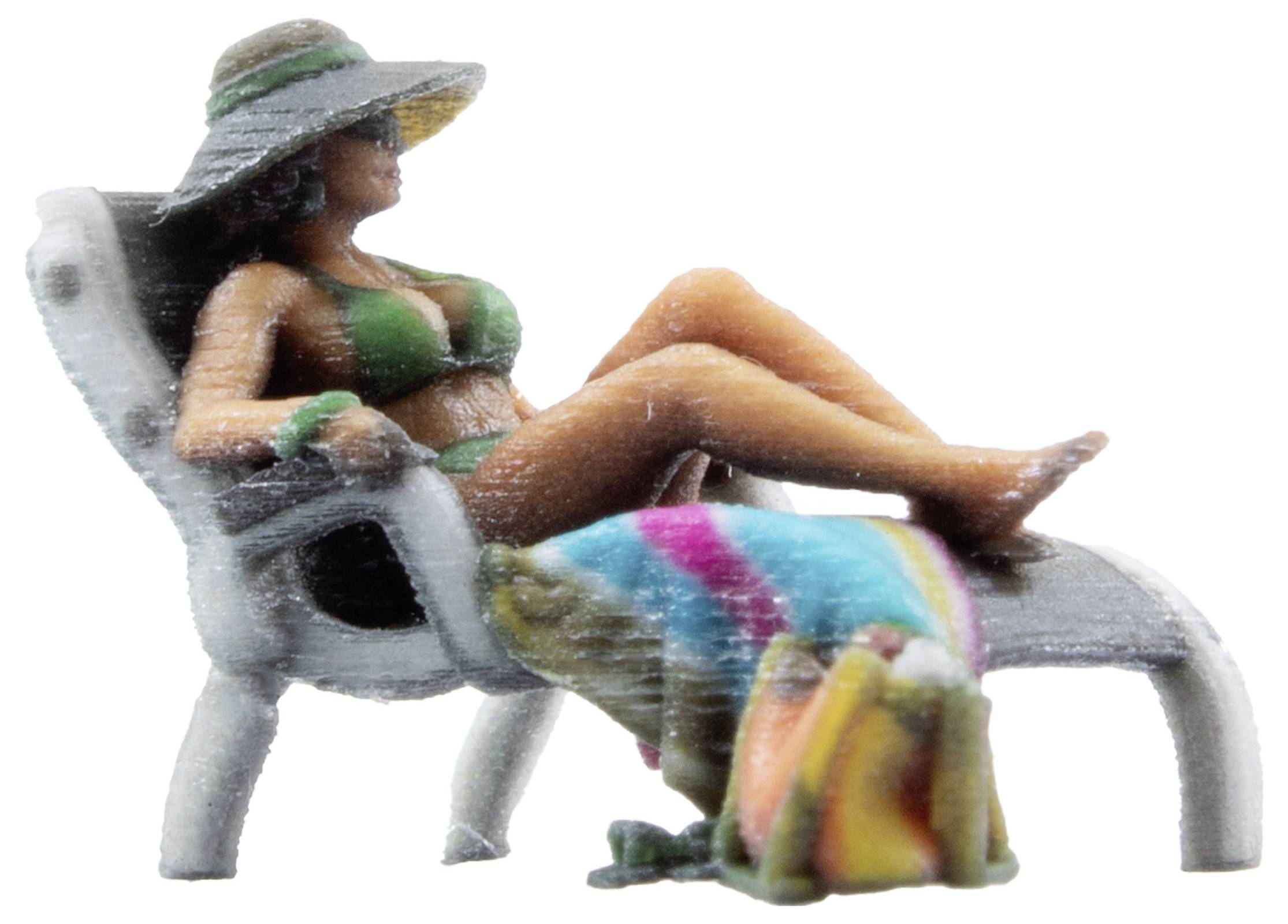 Une figurine miniature d'une femme en bikini vert est allongée de manière détendue sur un transat, portant un chapeau et une serviette colorée.