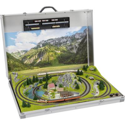 Petit système de train miniature dans une valise avec une scène de paysage : prairies, montagnes, lac, arbres et trains sur des rails au premier plan.