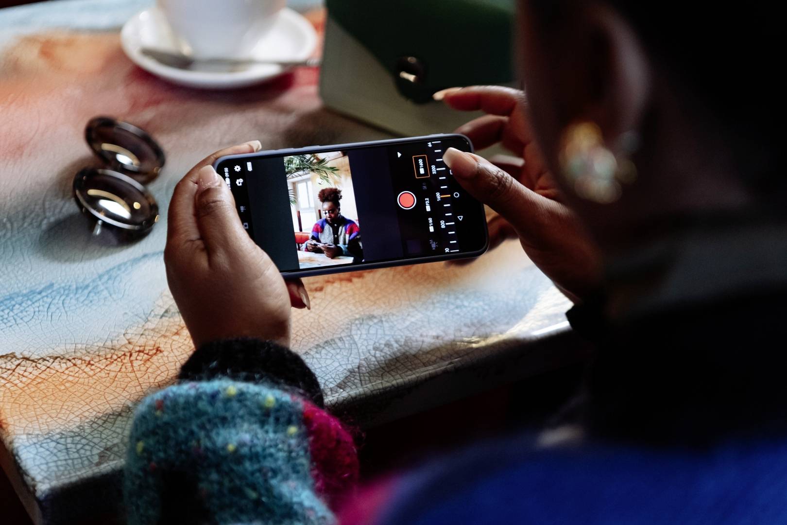 Une personne tient un smartphone et prend une photo dans un café. Des lunettes de soleil et une tasse de café sont posées sur la table.