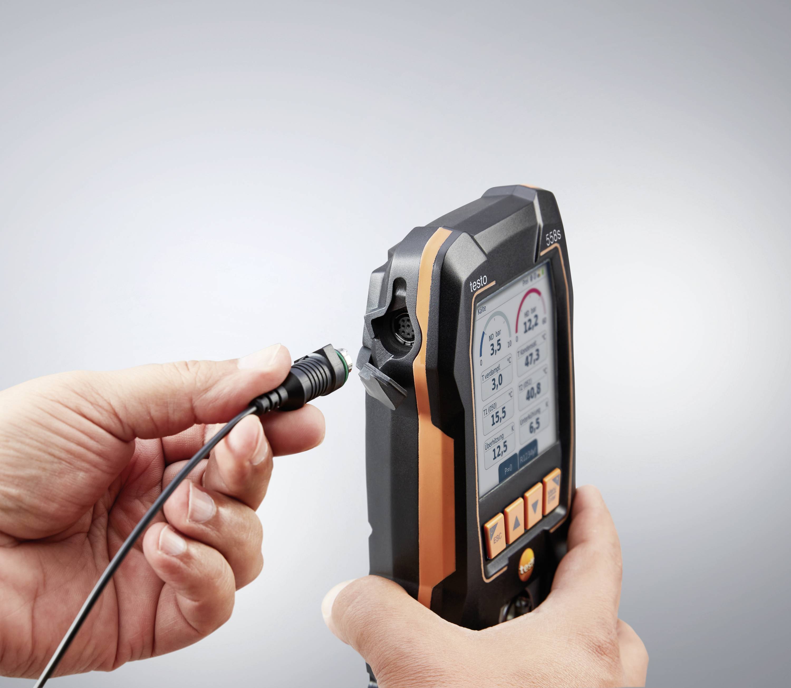 Une main tient un appareil de mesure portable avec un écran. Une deuxième main branche un câble dans l'appareil. L'écran affiche des valeurs de mesure.