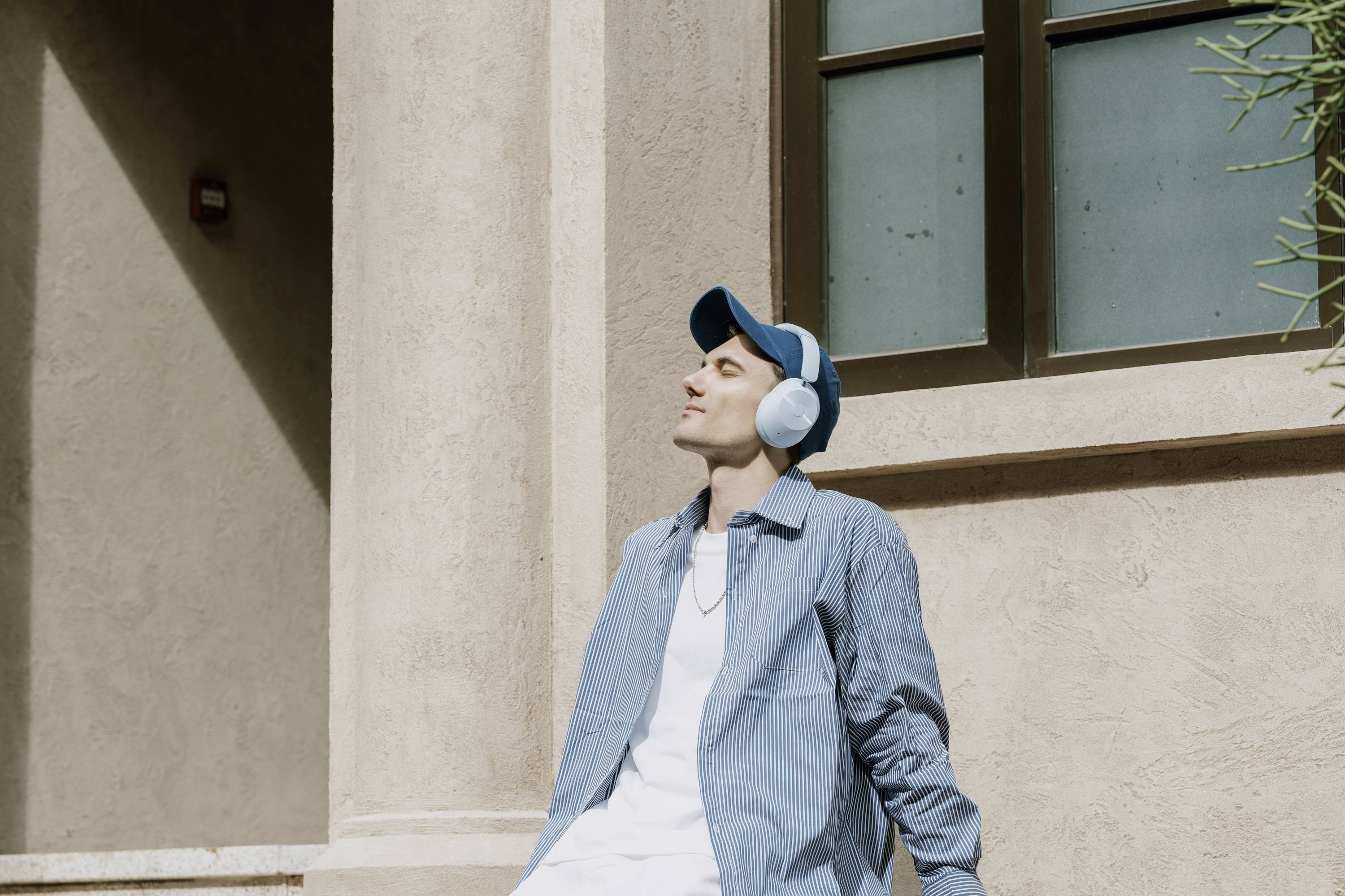 Une personne en tenue décontractée est assise dehors au soleil, profitant d'un moment de détente. Elle porte des écouteurs et une casquette bleue.