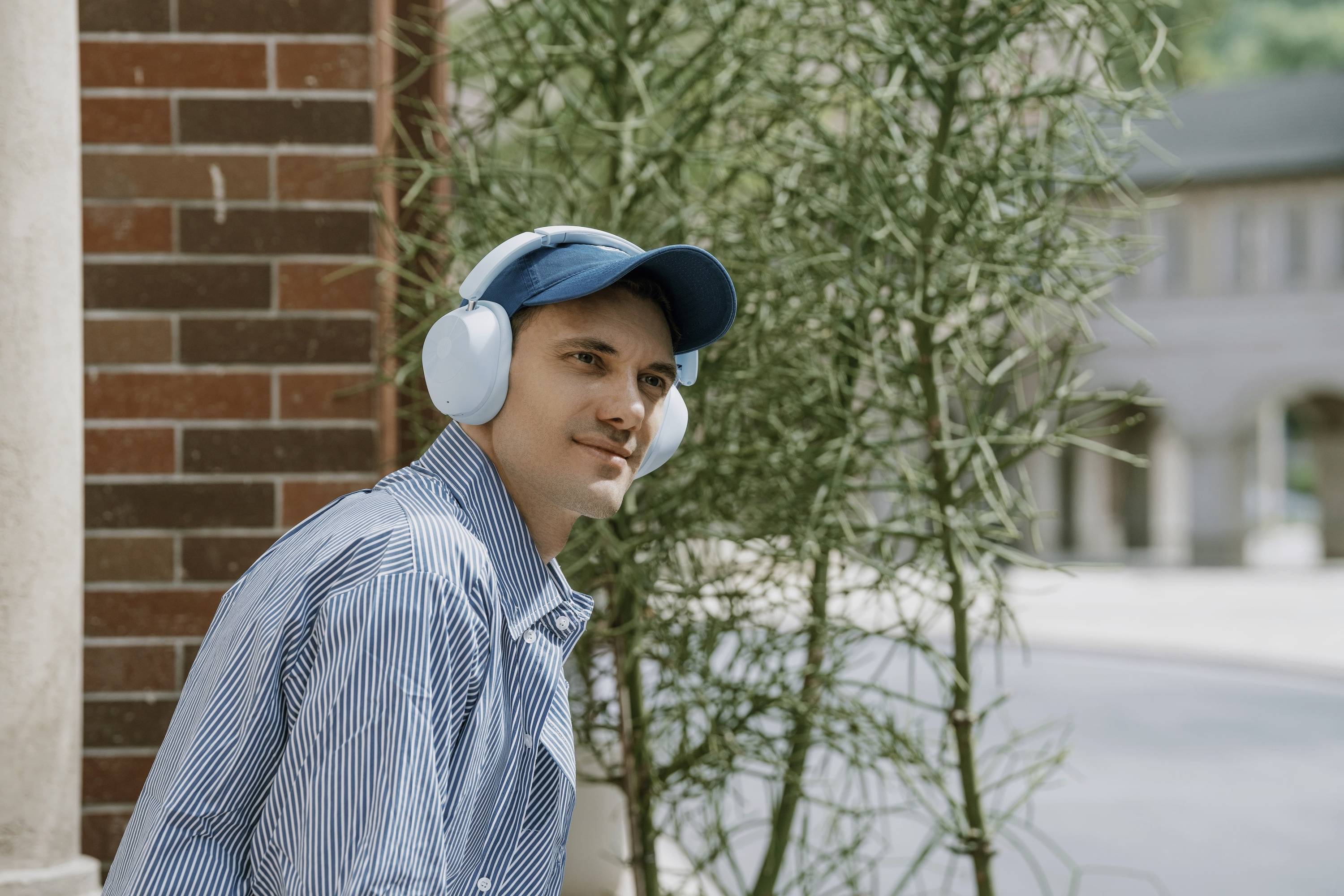 Un homme portant une casquette bleue et des écouteurs est assis dehors près d'une plante, regardant sur le côté, avec des bâtiments visibles en arrière-plan.
