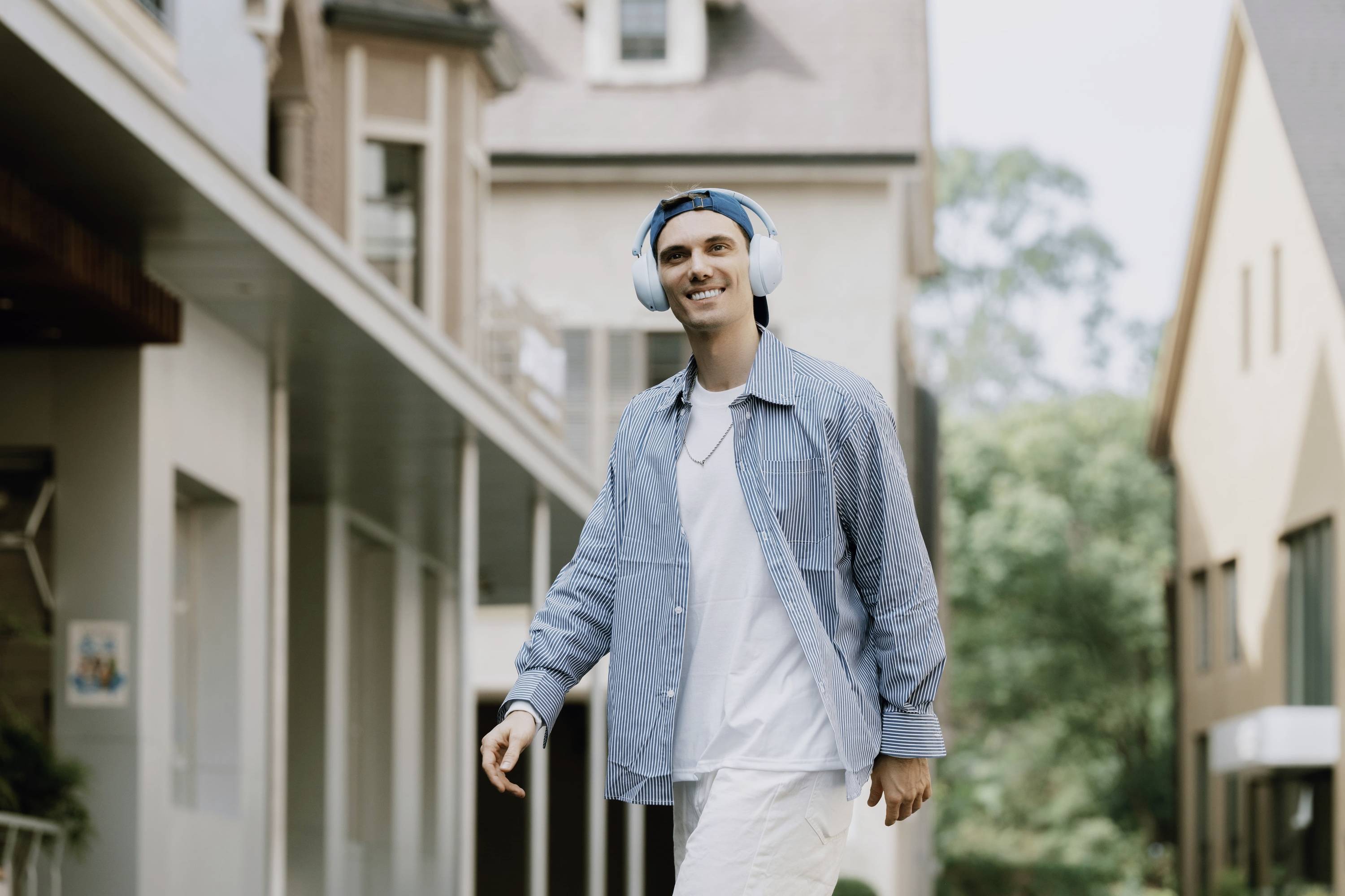 Un homme souriant portant des écouteurs marche devant des bâtiments modernes. Il porte une chemise bleue et un pantalon blanc. Ambiance : détendue.