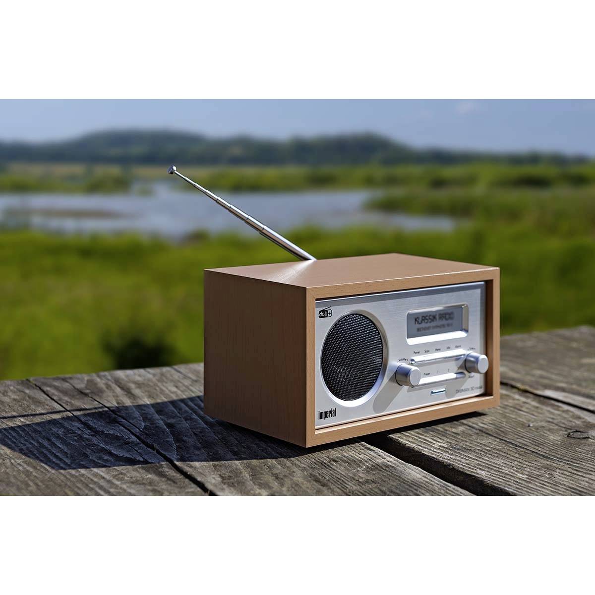 Un poste de radio rétro en bois est posé sur une table en bois à l'extérieur. En arrière-plan, un paysage de prairie et des collines apparaissent de manière floue.