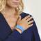 Une femme montre un bracelet bleu à son poignet, porte un chemisier bleu marine et a des cheveux blonds.