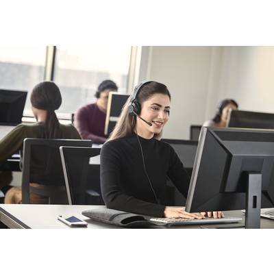 Une femme portant un pull noir et un casque travaille sur un ordinateur dans un bureau moderne avec d'autres personnes.