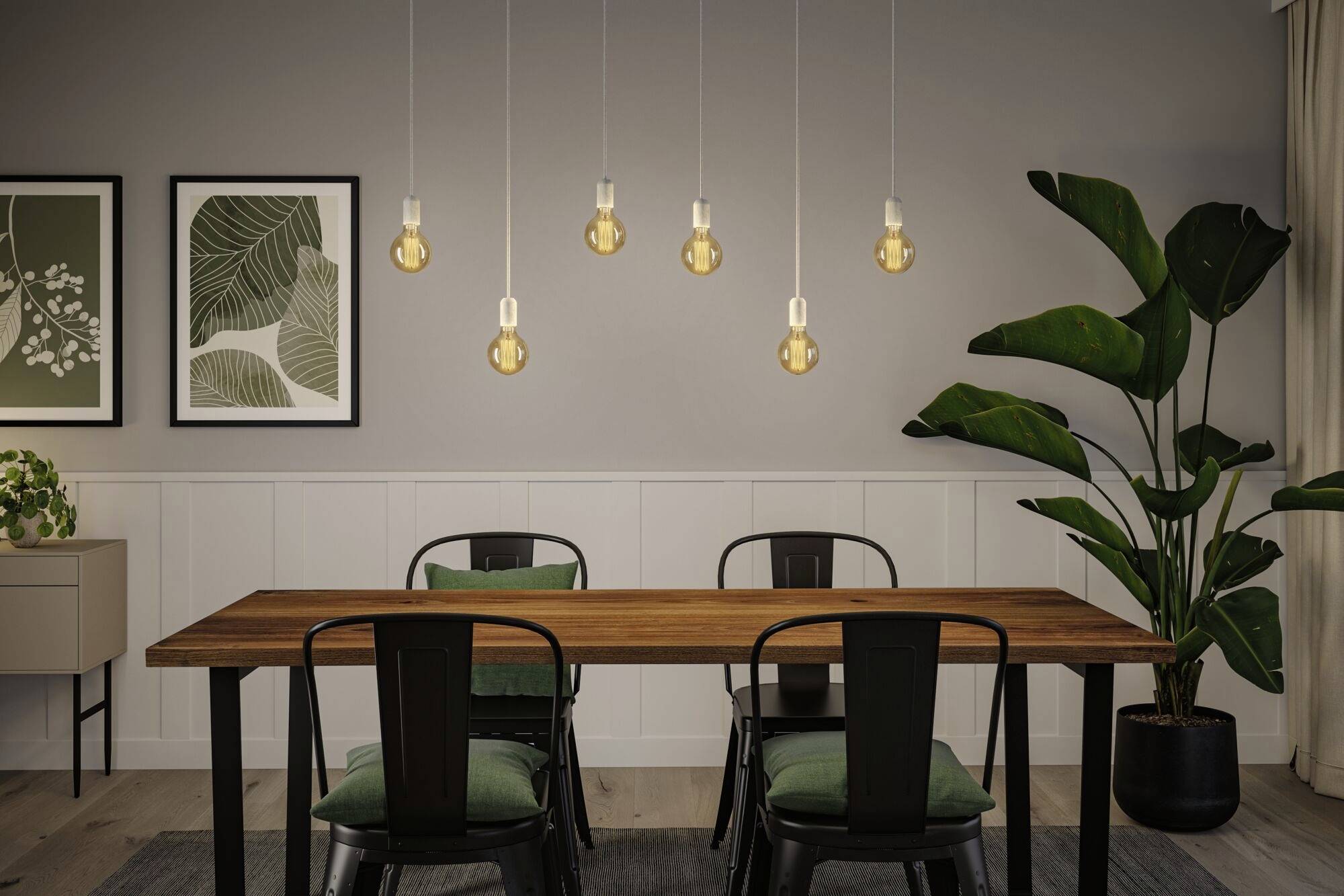 Une salle à manger avec une table en bois et quatre chaises. Six ampoules suspendues éclairent la pièce. Deux tableaux de plantes sont accrochés au mur.