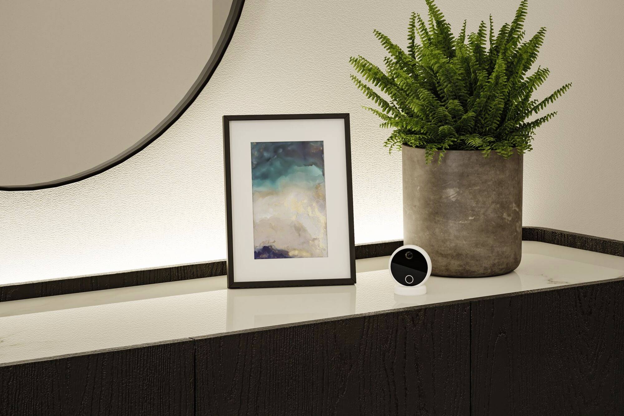 Œuvre encadrée et miroir rond sur un buffet sombre, à côté d'un grand fougère verte dans un cache-pot en pierre.