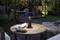 Une table ronde en bois dans le jardin avec une lampe noire, un livre, une tasse et un biscuit. À l'arrière-plan, on aperçoit un éclairage de jardin nocturne.