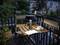 Une table en bois sur un balcon avec une lampe, un livre ouvert et une tasse de thé ou de café dans une scène de jardin paisible en fin de soirée.