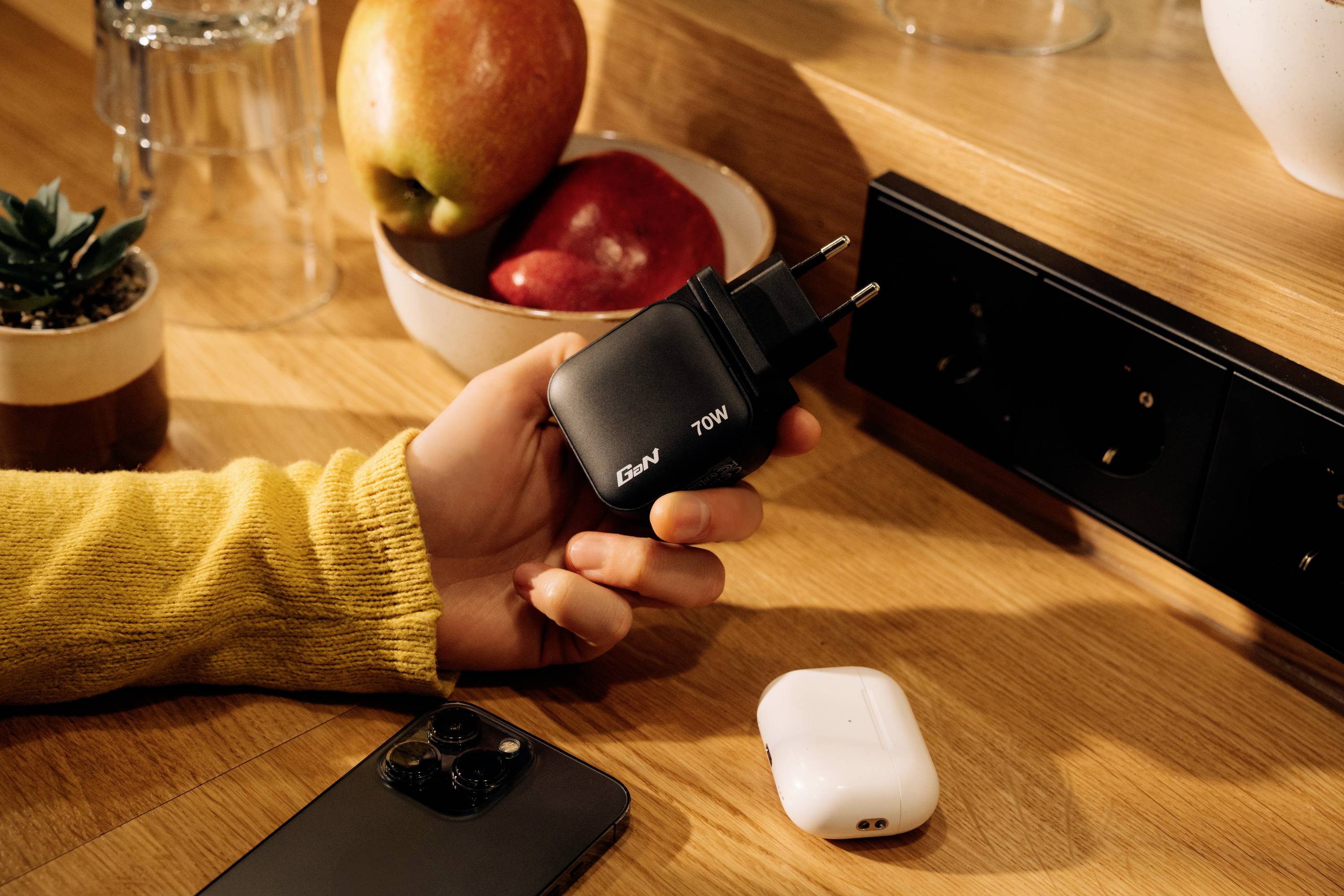 Une main tient un chargeur noir de 70W sur une surface en bois. À côté se trouvent un smartphone, des écouteurs sans fil et un bol de pommes.