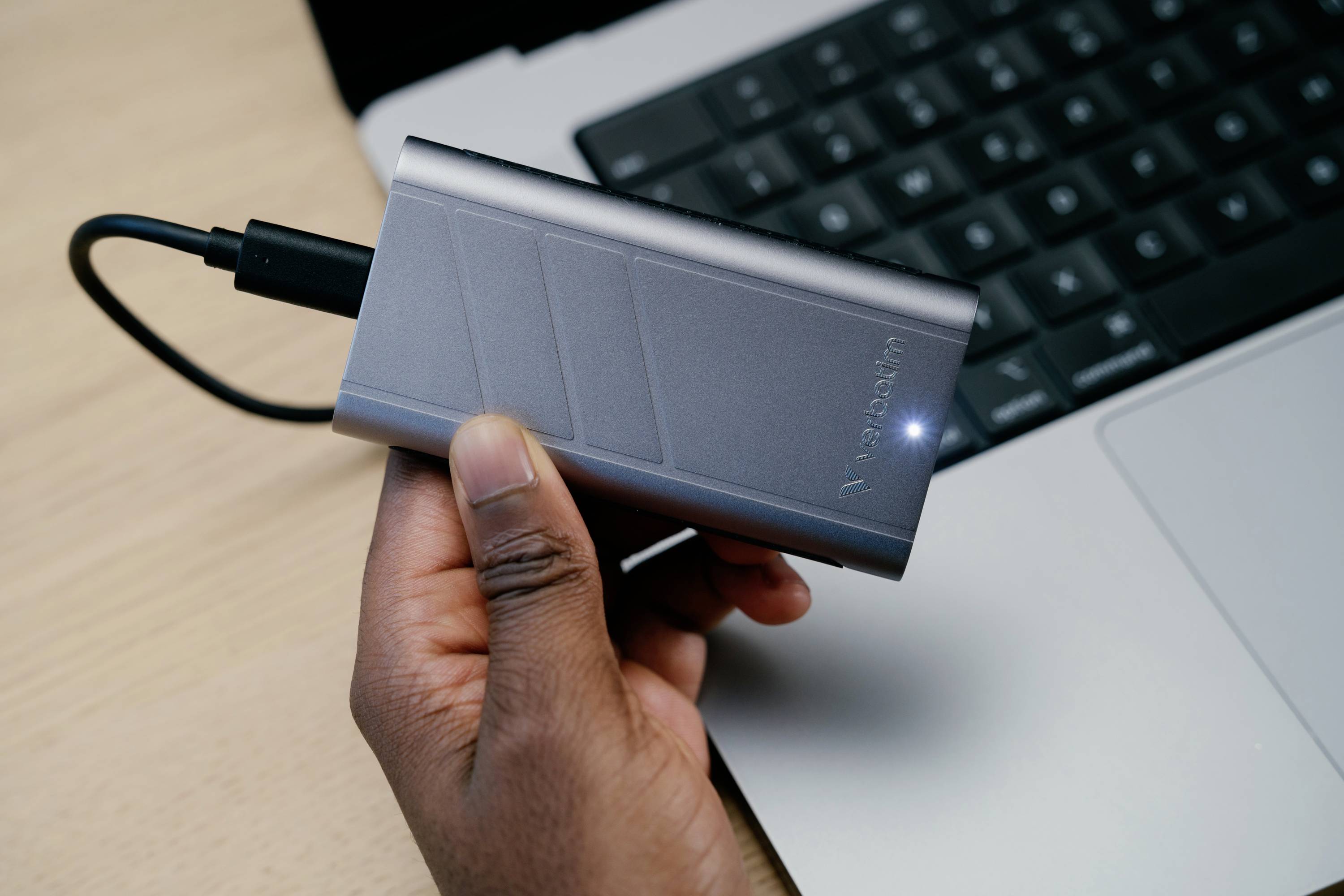 Une main tient un disque dur externe connecté par un câble à un ordinateur portable. Le portable dispose d'un clavier noir.