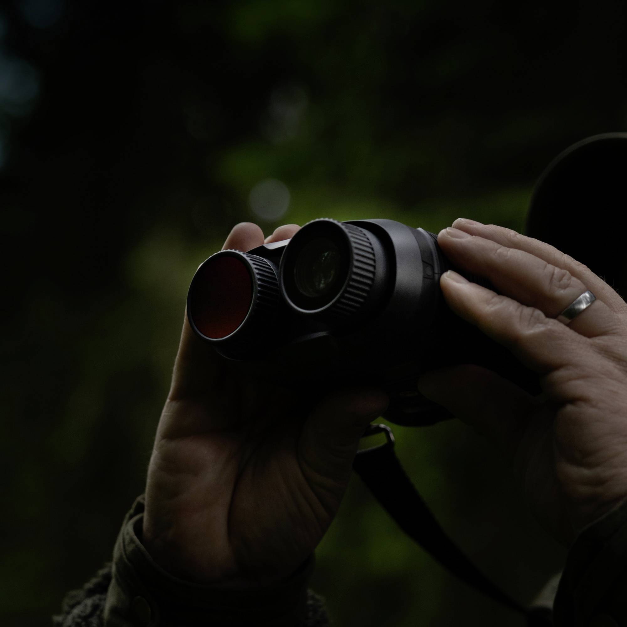 Une personne tient des jumelles à lentilles rouges, prête à observer. L'éclairage est tamisé, suggérant un environnement naturel.
