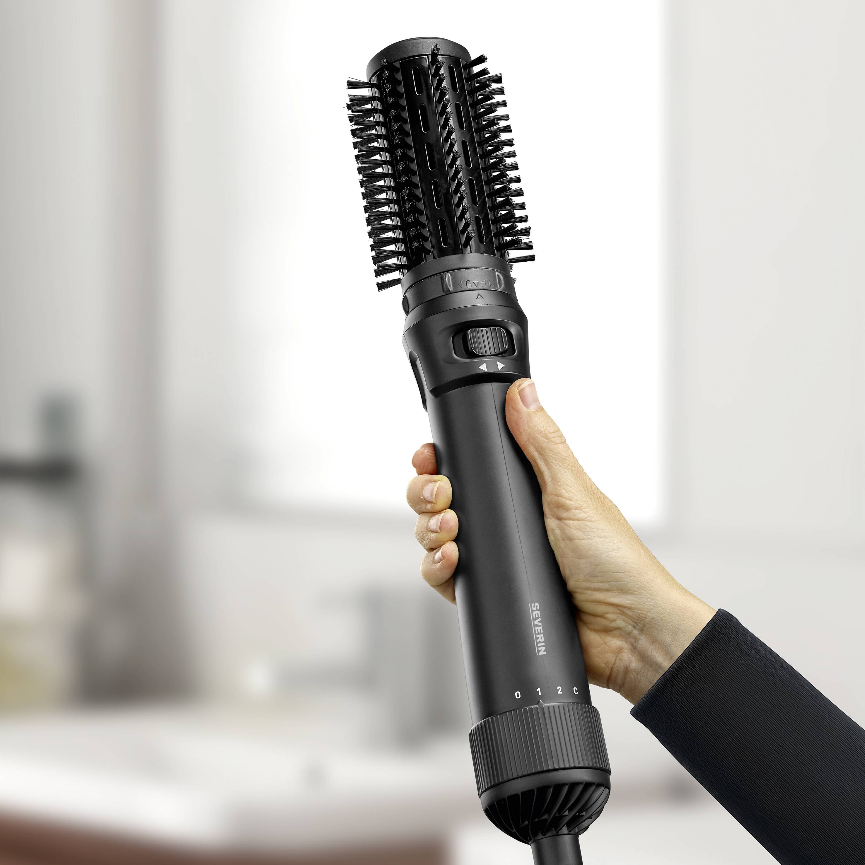 Une main tient une brosse à air chaud noire avec une tête de brosse rotative, adaptée pour le coiffage des cheveux. En arrière-plan, flou.