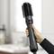 Une main tient une brosse à air chaud noire avec une tête de brosse rotative, adaptée pour le coiffage des cheveux. En arrière-plan, flou.