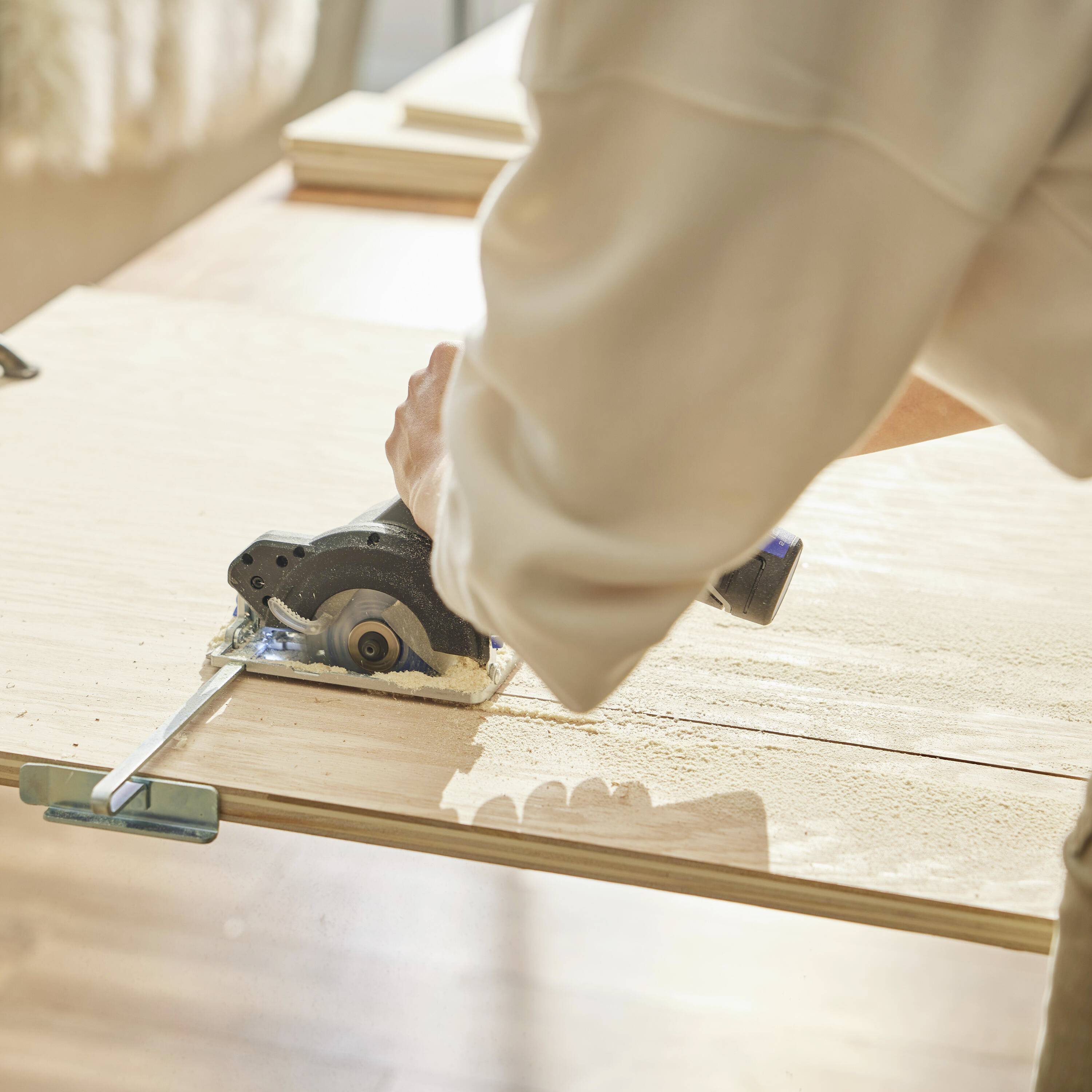 Une personne utilise une scie circulaire manuelle pour couper une planche en bois. La scène montre un espace de travail avec une table et une lumière vive.