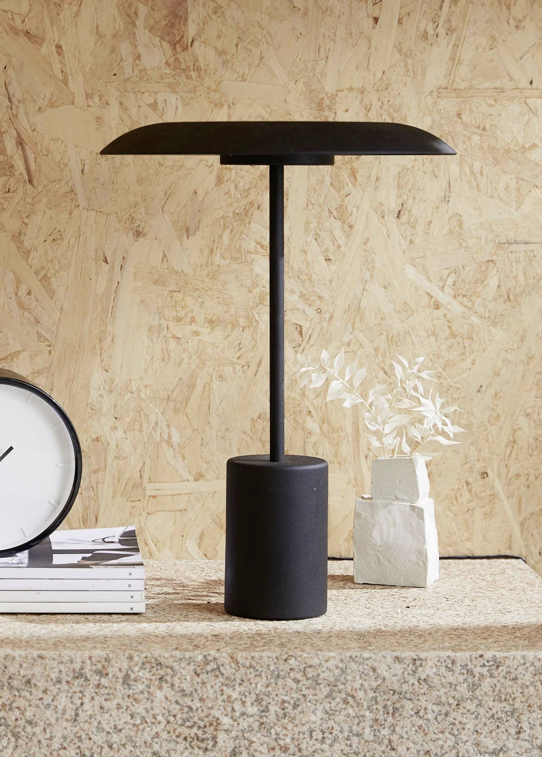 Lampe de table noire à socle cylindrique posée sur une table, à côté d'une pile de livres, une horloge et une petite plante.