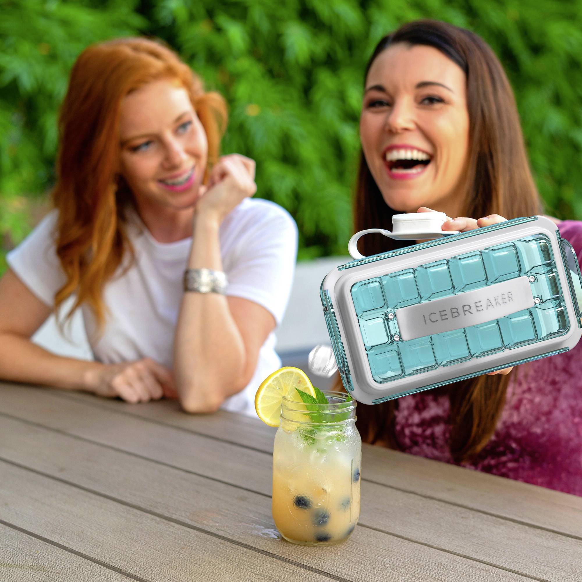 Deux femmes rient assises à une table à l'extérieur. Au premier plan se trouve une boisson avec une lime. L'une des femmes tient un bac à glaçons vert.