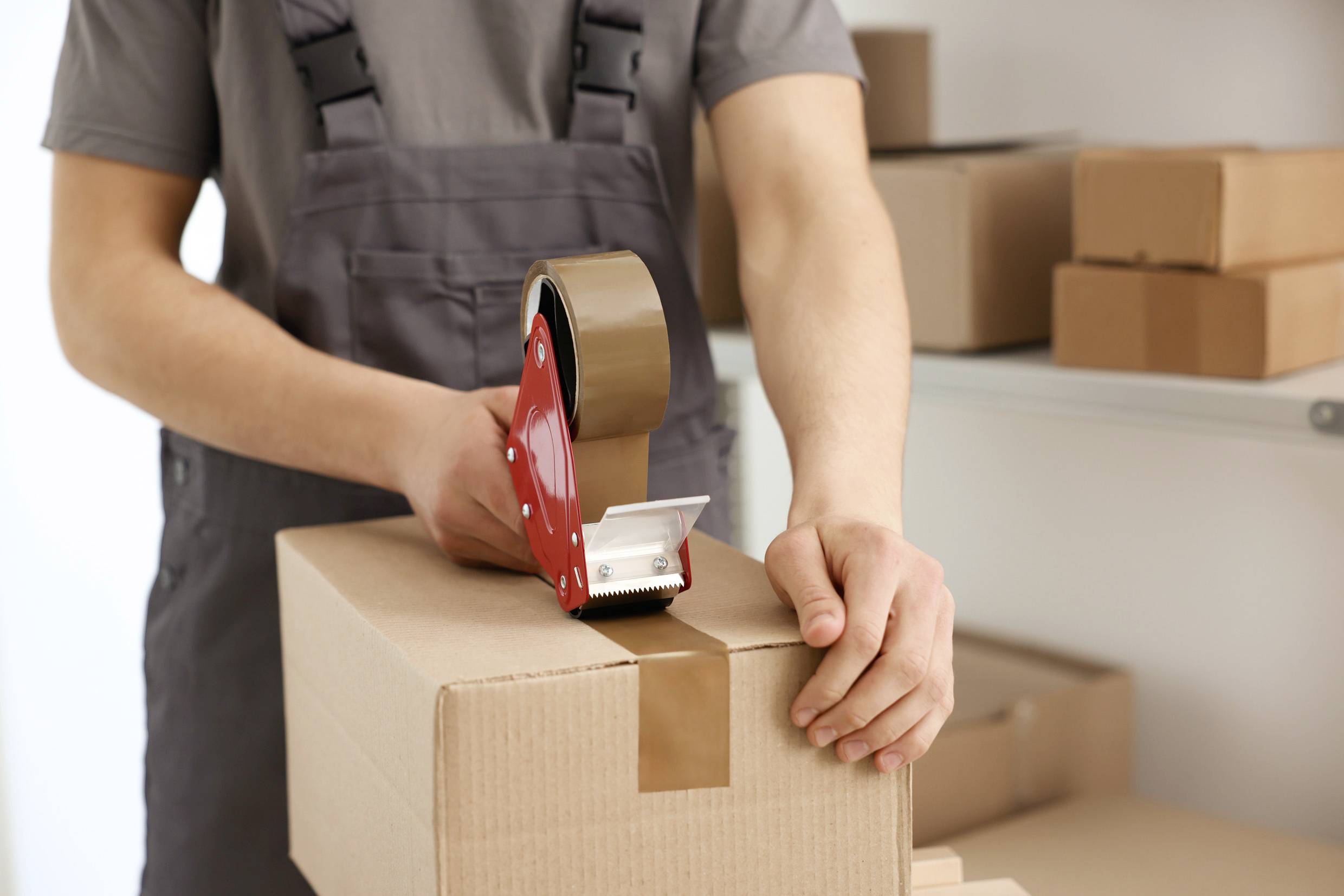 Une personne emballe un carton avec un dévidoir de ruban adhésif. En arrière-plan, on peut voir d'autres cartons sur une étagère.