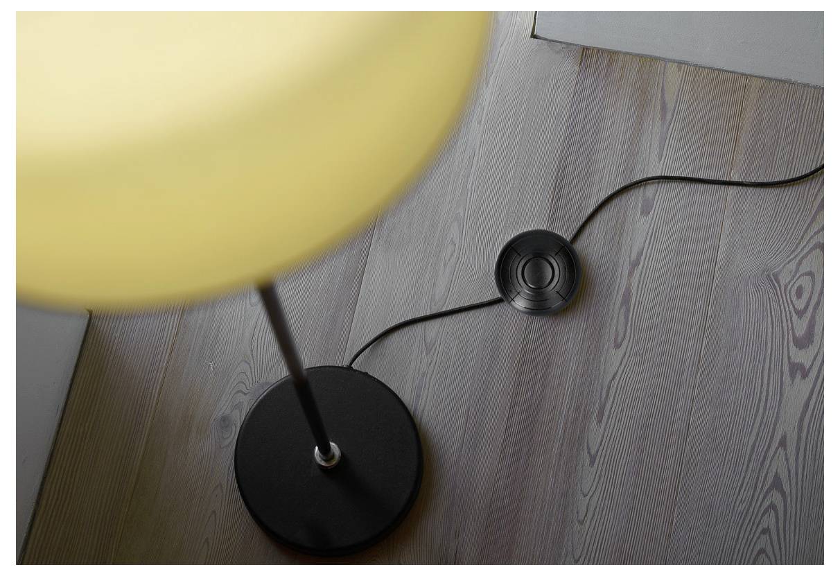 Lampadaire avec interrupteur rond noir sur un sol en bois brun clair. L'abat-jour est représenté de manière floue et brille d'une lumière jaune pâle.