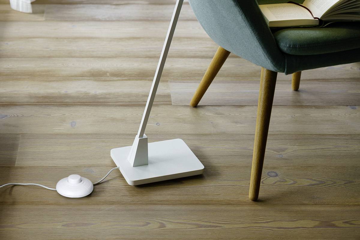 Une lampe moderne et minimaliste au socle carré posée sur un parquet, à côté d'un fauteuil vert sur lequel repose un livre ouvert.