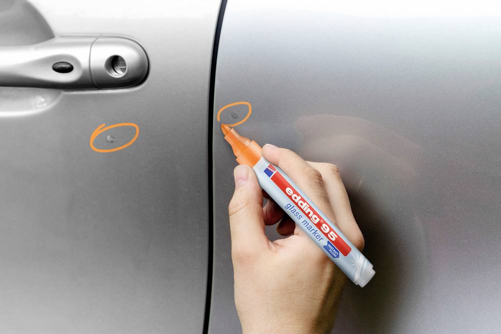 Une main marquant deux rayures sur une portière de voiture avec un stylo orange pour les réparer.