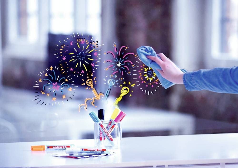Une main essuie un dessin de feu d'artifice sur une surface en verre. Au premier plan, des marqueurs de tableau blanc colorés sont disposés dans un gobelet.