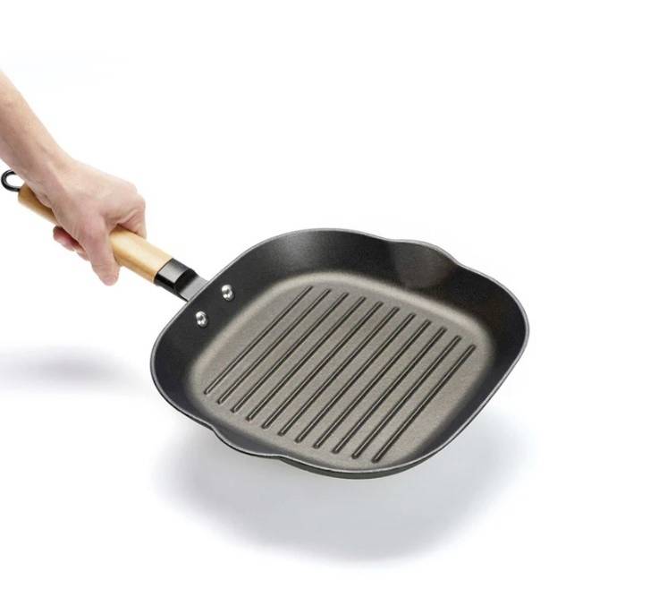 Une main tient une poêle à griller carrée avec un manche en bois, comportant des rainures pour créer des marques de grill. Elle est noire et en métal.