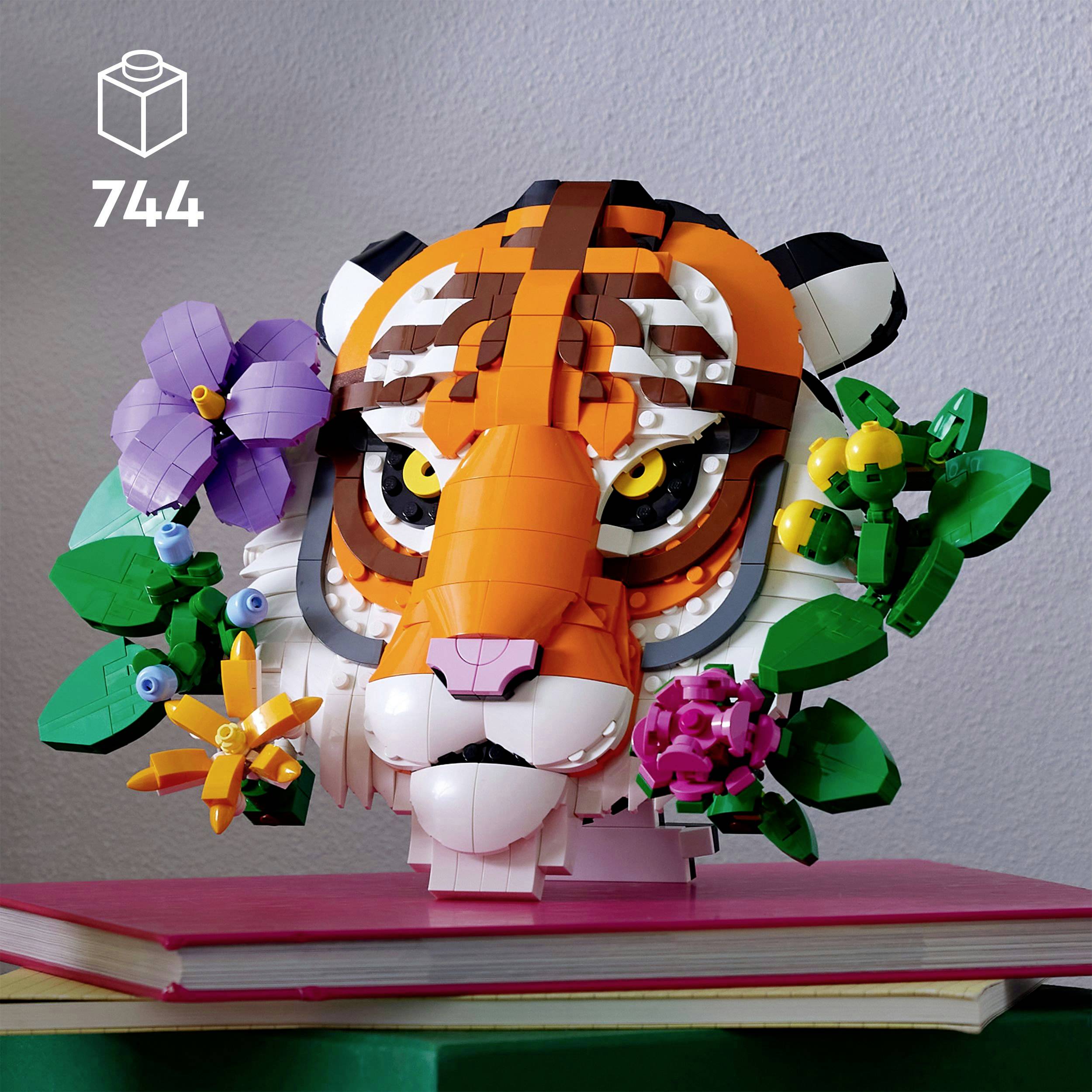 Une tête de tigre en Lego avec des fleurs et des feuilles, posée sur un livre rouge, devant un arrière-plan uni.