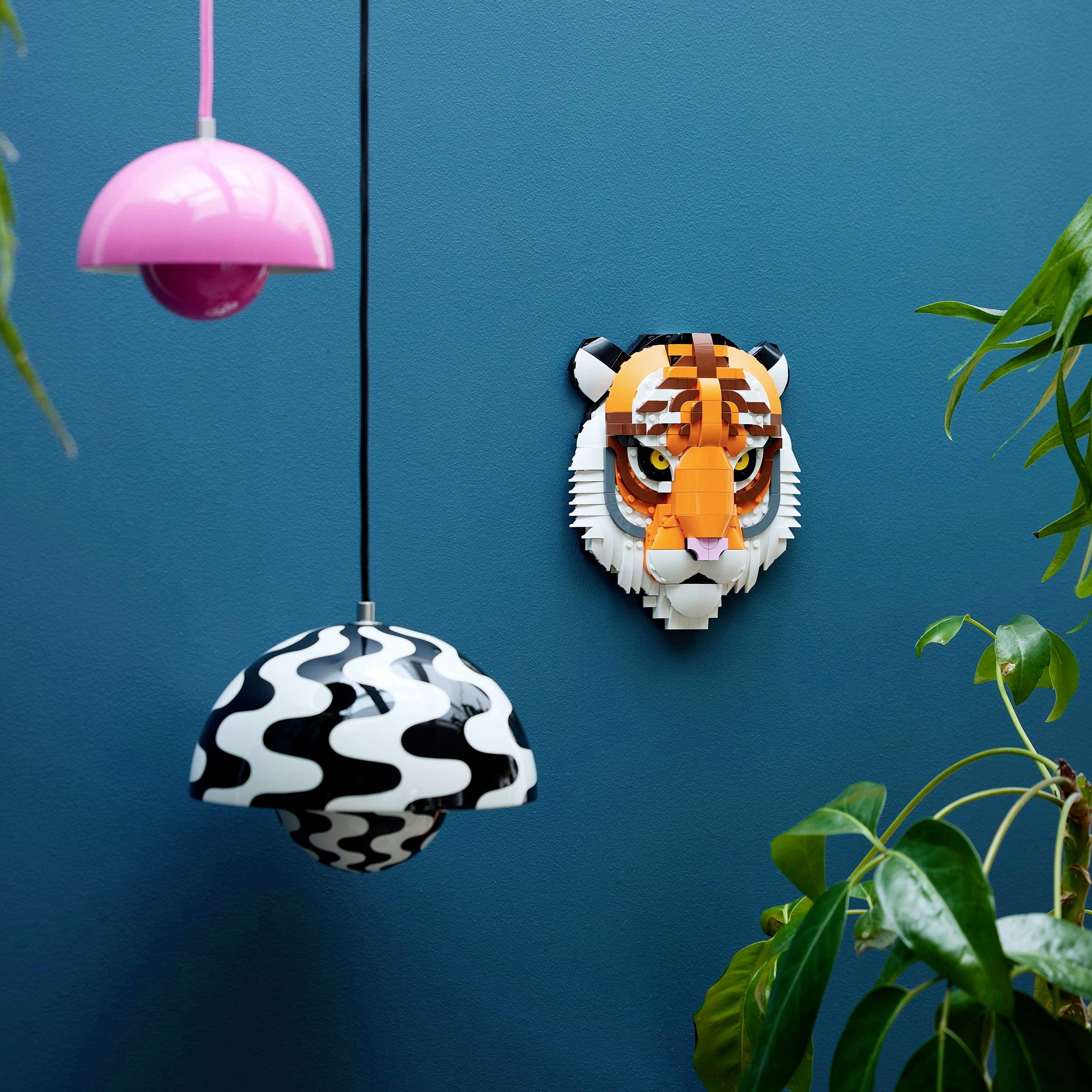 Une tête de tigre en Lego est accrochée à un mur bleu. À côté, on peut voir des lampes colorées et des plantes.