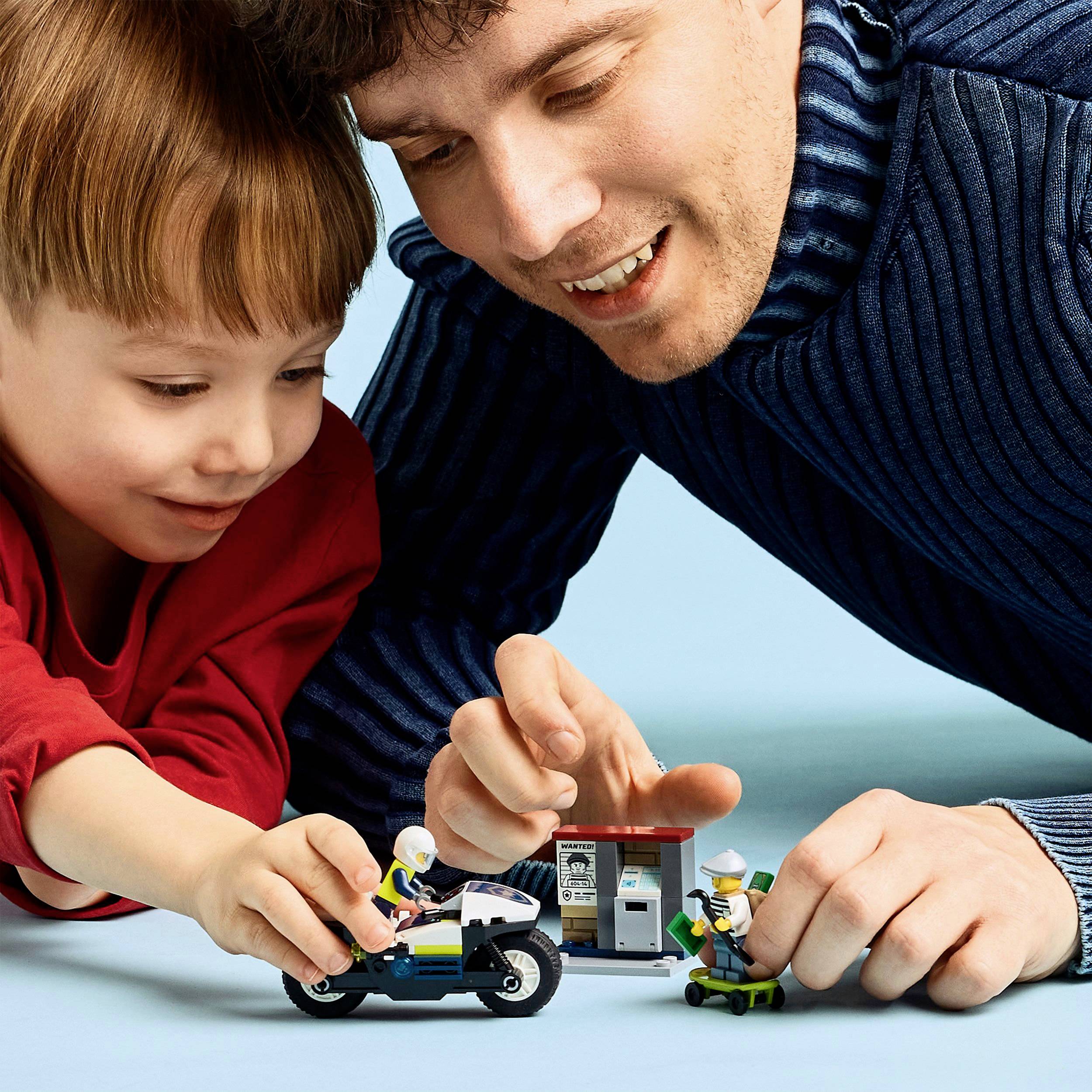 Un homme et un enfant jouent avec un set Lego de moto sur une table bleue. Ils sont joyeux et concentrés.